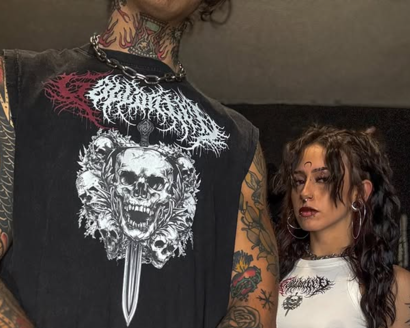 Two people with tattoos standing close together, one wearing a black sleeveless shirt with a skull design.